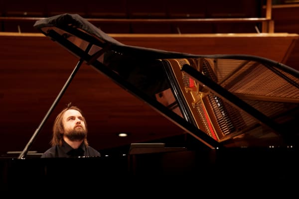 Daniil Trifonov playing with the NZSO. Daniil is sitting at a grand piano, he has shoulder length brown hair and a beard, and is wearing a black shirt.