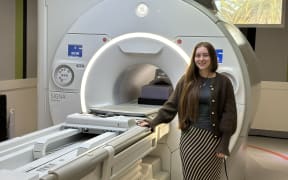 Taylor stands to the right of the MRI machine with her right had resting on it. She has waist length hair worn down over one shoulder, with a stripy long black and white skirt, green t-shirt and brown cardigan. The machine towers over her, with the circular entrance for patients lit up.