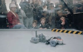 A group of school children watch two robots fighting behind a glass wall.