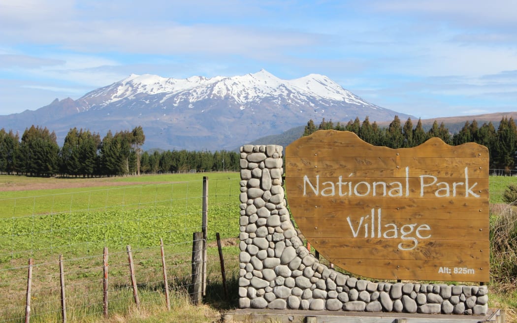 A sign at the entrance to National Park Village that says 'National Park Village', with Mount Ruapēhu visible in the background.