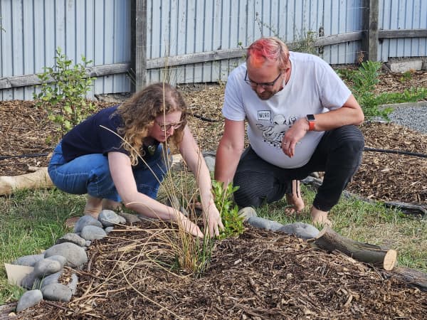A woman and a man are shown squatting in a patch of garden. She is parting the leaves of a plant. The garden is bordered by stones and contains a native plant on a base of wood chips.
