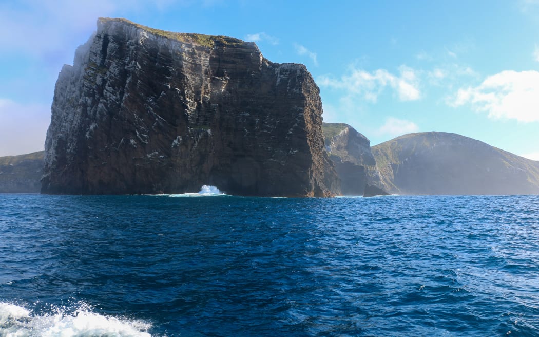 The sheer rocky cliff face of a rugged island rises out of deep blue ocean beneath a blue sky dotted with puffs of white cloud. The top of the island has only grassy/tussock vegetation.