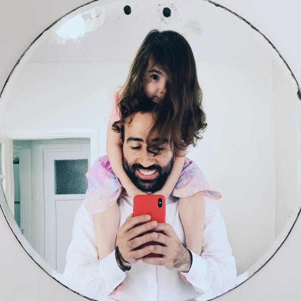 A bearded father takes a selfie with his daughter sitting on his shoulders.