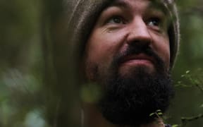 Sam Gibson, a bearded man wearing a beanie, looks to the sky with forest behind him.