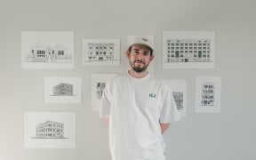 Josh stands in front of a white wall with prints of his illustrations tacked on to it.