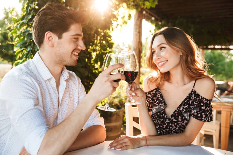 Picture of young loving couple sitting in cafe by dating outdors in park holding glasses of wine drinking.
