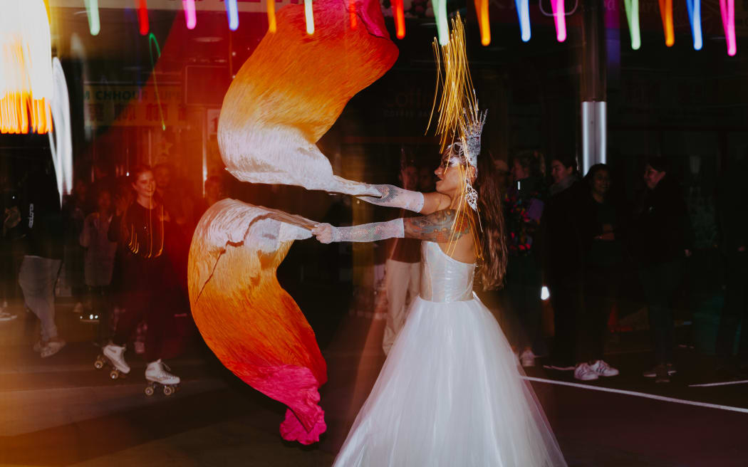 A person in a white dress is dancing in the street waving some orange scarves. It is early evening. There is someone roller skating in the background, and a crowd of people watching.