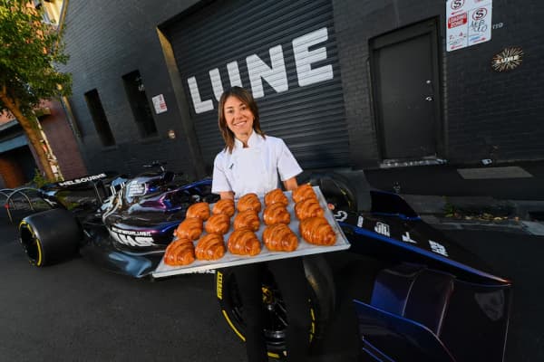 A brown-haired woman in a white shirt holds a tray of cooked croissants.