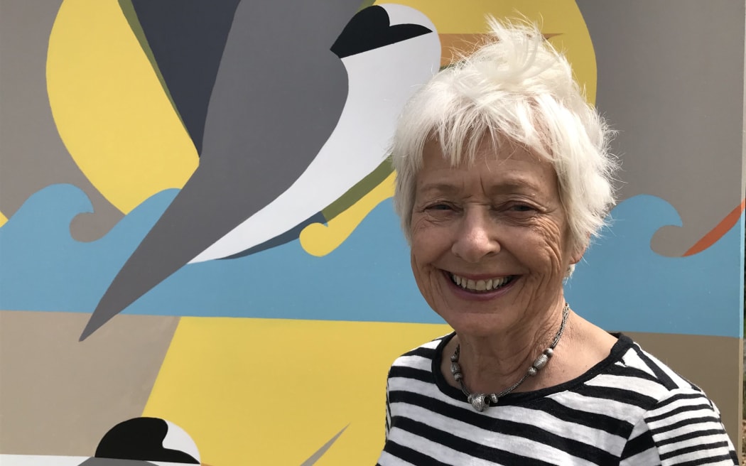 New Zealand Fairy Tern Trust convenor Heather Rogan smiling in front of a painted mural of a fairy tern. She has short white hair, a striped shirt and a necklace.