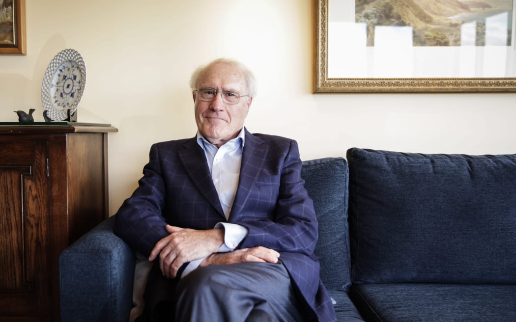 Geoffrey Palmer sits on a couch and looks at the camera.