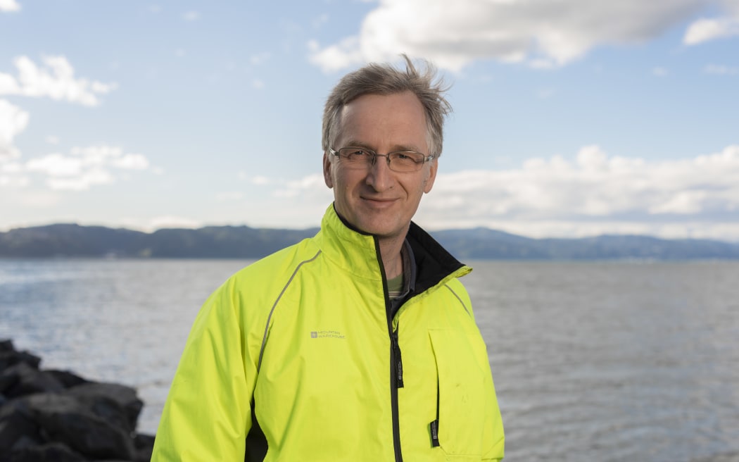 A man in a fluoro yellow zip-up jacket stands in front of a body of water. He is wearing glasses, smiling, and his hair is being lifted by a breeze.