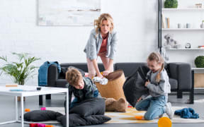 A photo of an emotional tired mother putting toys in basket while naughty kids fighting with pillows