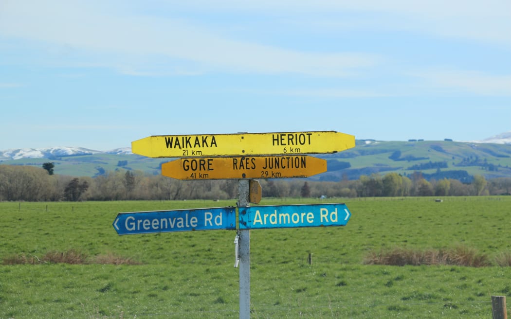 At a junction - a street sign shows 41 kilometres to Gore or 6 kilometres to Heriot with pastures and rolling hills laying behind