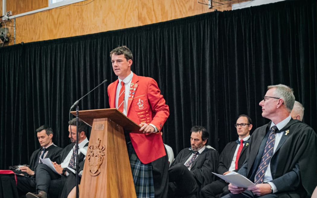 Fergus Morunga is dressed in a red blazer delivering a speech behind a podium.