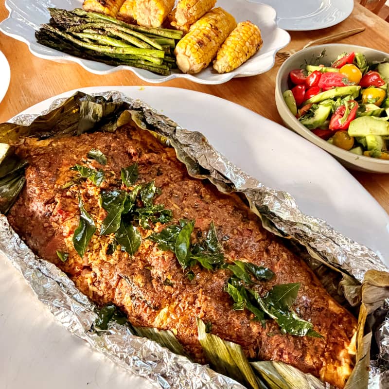 A dish of coconut chilli barbecue salmon on a bed of foil, with asparagus and sweetcorn on dishes in the background.