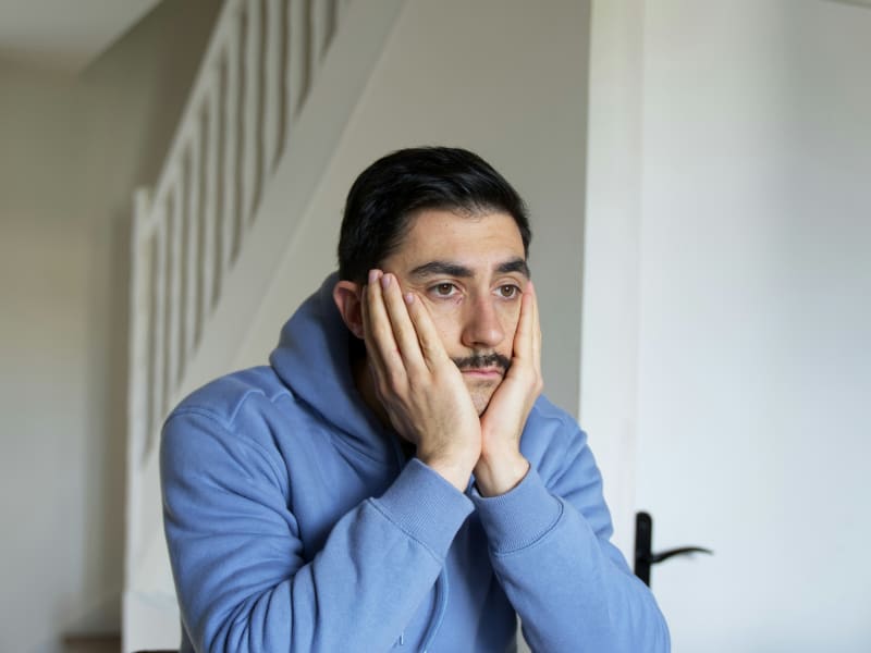 A grumpy man with a moustache and his head in his hands looks into space.