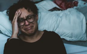 A woman with short hair and black-rimmed glasses cries on the edge of an unmade bed.