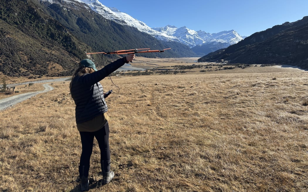 A woman in a black puffer jacket stands on the dry brown grass of the valley floor, facing away from the camera, her arm extended up high, holding a radio antenna skywards. There is a gravel road snaking along the left of the valley, next to the steep green mountain slopes that extend up to snowy peaks and a bluebird sky. It looks crisp but cool.
