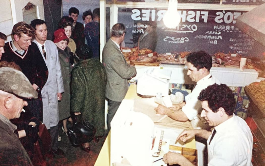 This is an old photo of a queue of people inside Theo's fish and chip shop in Christchurch.. The queue is out the door. The photo is grainy and clearly taken on film. I'd guess it's sometime around the 70s.