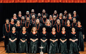 Pacific Connection Choir pose as a group. Their winning trophy is in the centre, on the floor in front of the group.