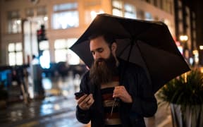 Mid adult man walking in city at night, using umbrella, looking at smartphone, Downtown, San Francisco, California, USA (Photo by Wonwoo Lee / Image Source / Image Source via AFP)