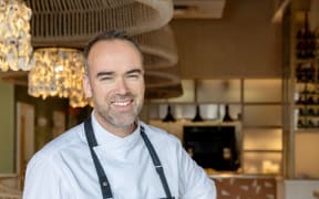 Toby stands in his restaurant wearing an apron over a white chef's shirt. He smiles.