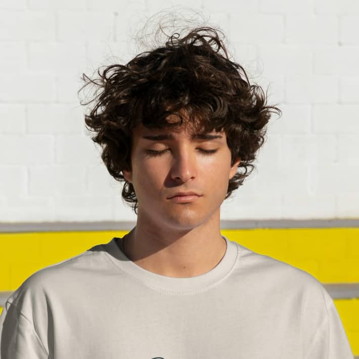 A boy with curly dark hair closes his eyes and stands against a white wall.