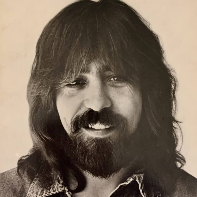 Clarence White, with long hair and beard, smiles and looks to the side.