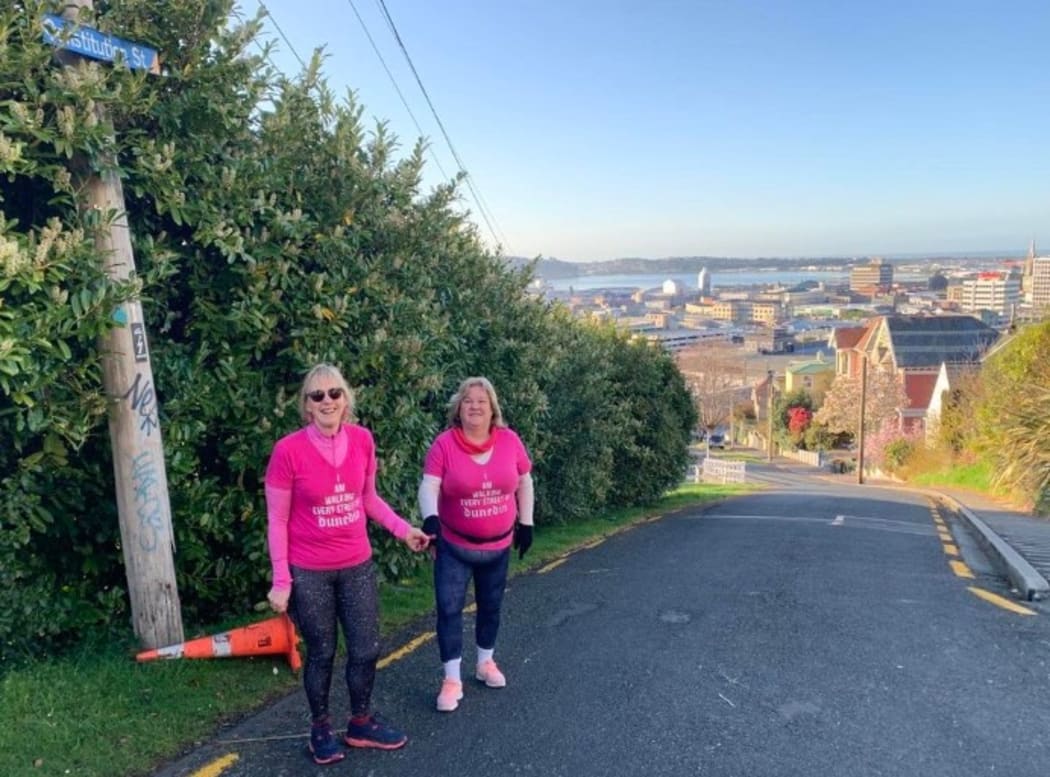 Julie Woods with her friend and sighted guide, Jo Stodart wearing pink t-shirts at the top of a steep Dunedin street.  The City can be seen in the background.