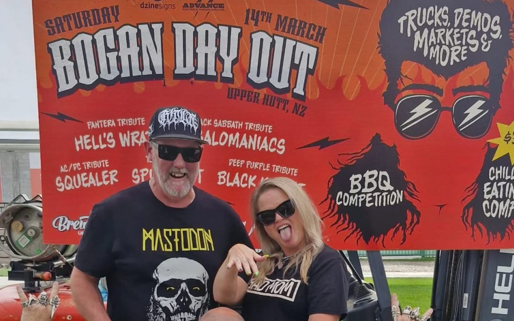 Bekz and Hayden stand in front a sign that reads "Bogan Day Out," They are both wearing black band t-shirts.