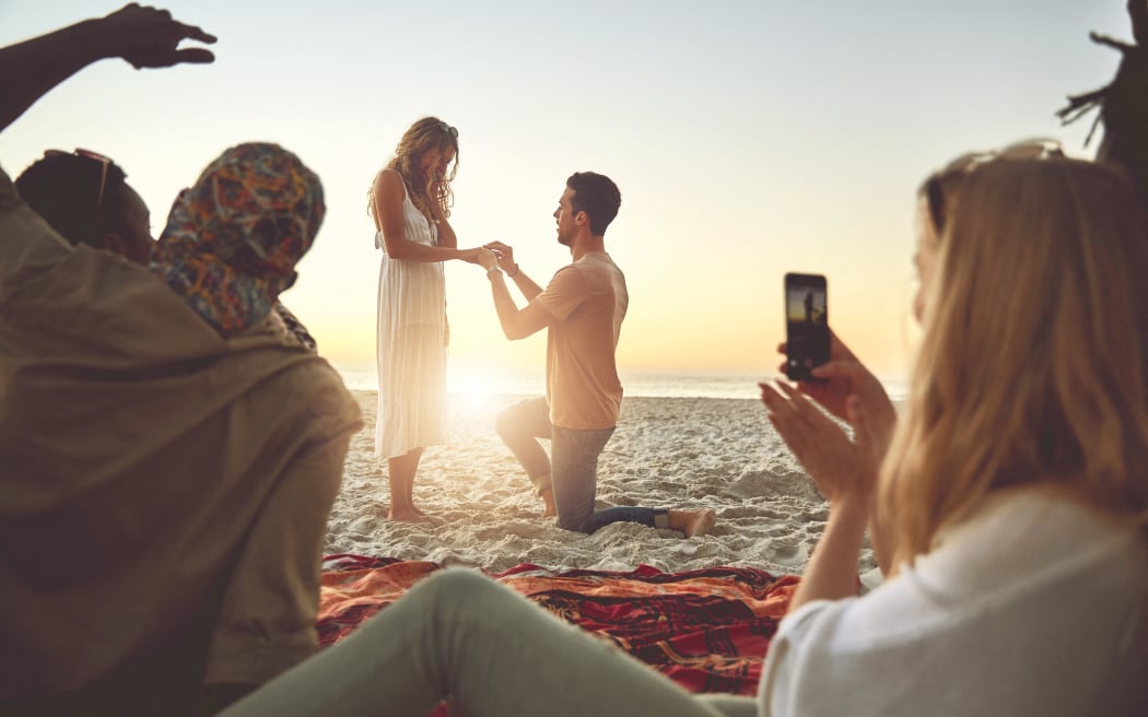 Young man proposing to woman on sunny summer beach with friends. (Photo by CAIA IMAGE/SCIENCE PHOTO LIBRARY / NEW / Science Photo Library via AFP)