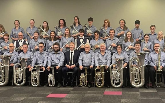 The Rolleston Brass pose for a band photo, seated in rows inside a large room. They all wear blue shirts and hold their instruments, smiling.