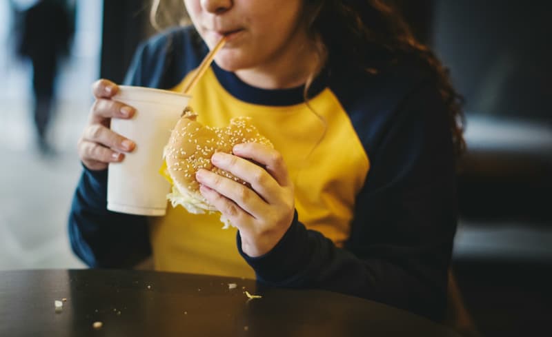 A young person sipping on a fast food drink in one hand and burger in the other.