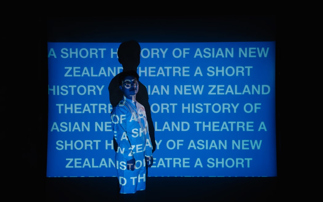 Nathan stands in front of a wall, wearing white, with a projector beaming a blue screen onto him, with the words "A SHORT HISTORY OF ASIAN NEW ZEALAND THEATRE" written in white, all-caps text over and over.