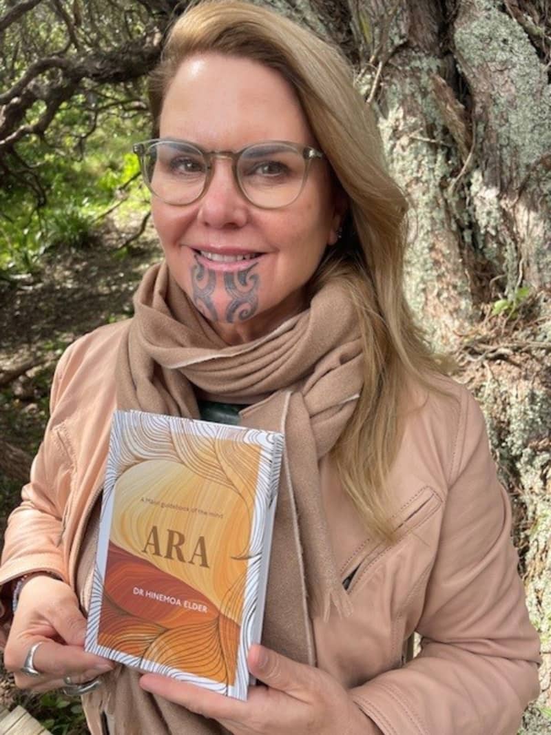 Dr Hinemoa Elder, wearing a brown scarf and brown jacket, holds a copy of her new book Ara.