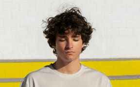 A boy with curly dark hair closes his eyes and stands against a white wall.