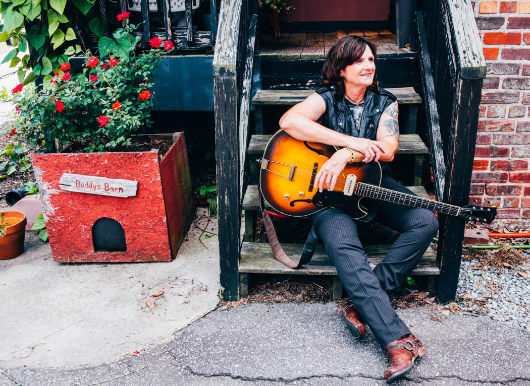 Singer-songwriter Amy Ray sits on outside steps with her guitar; red flowers in a planter box nearby.