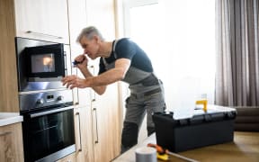 Aged repairman in uniform working, examining broken microwave in the kitchen using flashlight. Repair service concept. Selective focus.