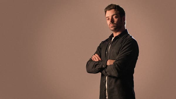 A man wearing a black shirt folds his arms and poses for a photo against a brown background.