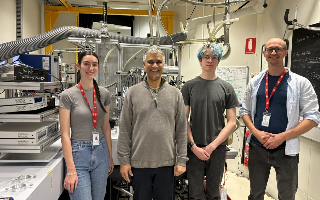 A woman and three men stand in a line in front of a large piece of metal scientific equipment with lots of pipes and tubes in a lab.