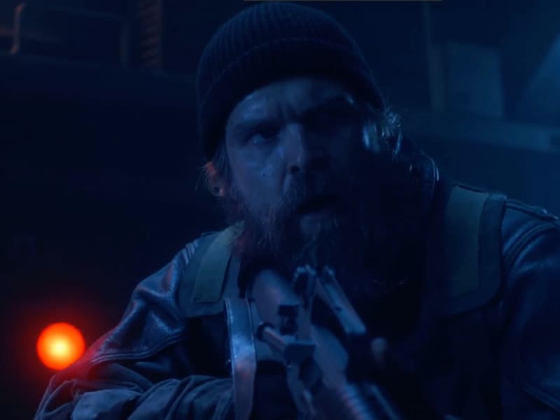 David Harbour, bearded and wearing a beanie, holds up a gun and looks nervous in low light.