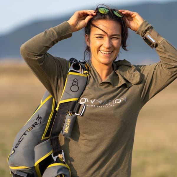Shayni Couch holds her sunglasses on her head and smiles widely while wearing a brown top and grey backpack.