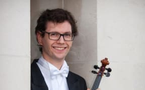 Alexander McFarlane is a viola player with the New Zealand Symphony Orchestra. He is wearing black and white concert attire, and holds his viola on his knee.