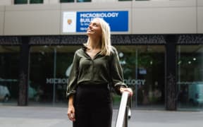 Jemma stands before the microbiology building in a green shirt and black pants. She's smiling, looking upwards to the right and holding on to a handrail.