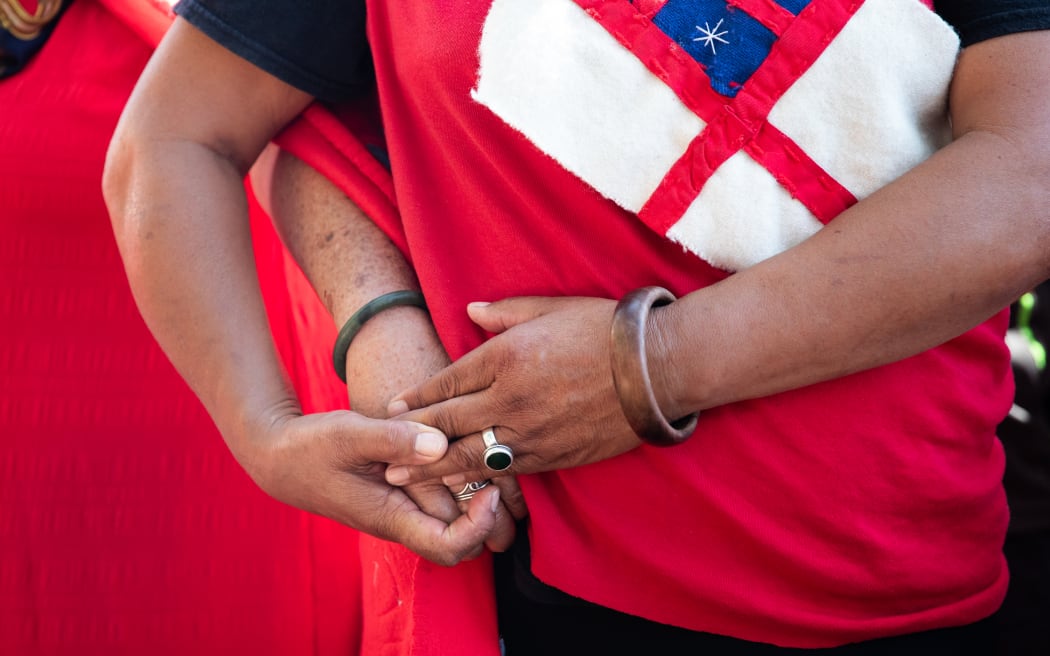 A close up of two wāhine Māori from Te Tai Tokerau holding hands as they stand with their backs turned on the Government in protest during Waitangi commemorations 2025. They are draped in homemade red woollen blankets hand-stitched with Tino Rangatiratanga and He Whakaputanga flags.