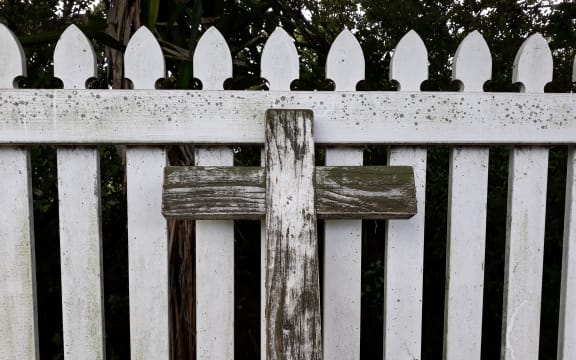 A wooden cross leans against a white picket fence.