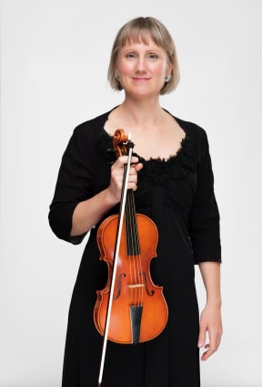 Musician Miranda Hutton stands holding her violin vertically.