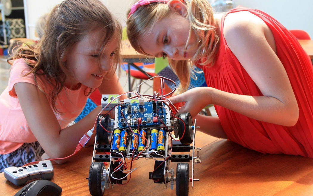 Two primary aged girls look at a robot on wheels on a  table
