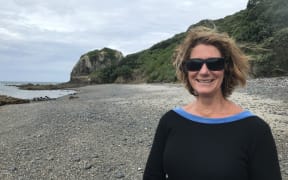 Sue is standing on a gravelly beach. She is smiling and wearing sunglasses.