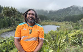 Mohi is standing at a point overlooking the Killarney lakes. The bigger lake is visible behind him with pine forest behind it. There's a lancewood tree directly behind him, and some harakeke and native trees visible around him. He's smiling, wearing a bright orange t-shirt. Both his hands are clasped together in front of him.